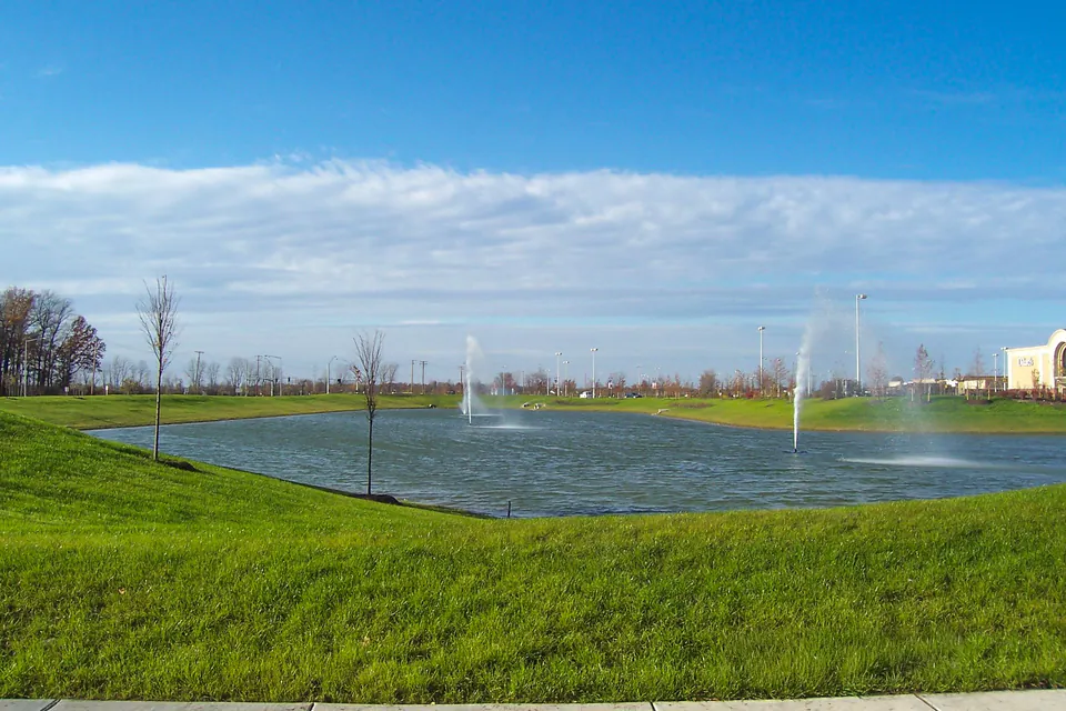 Commercial retention pond with fountains, mown lawn edges, sidewalk, and open sky